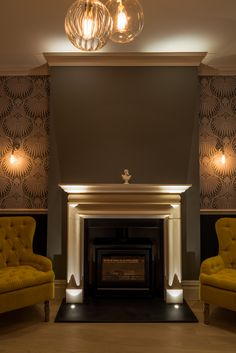 two yellow chairs sitting in front of a fire place with lights on the wall above it