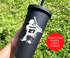 a hand holding a black tumbler cup with the number 27 on it and an ad for adams's