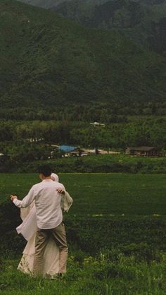 a man and woman walking through a lush green field with mountains in the back ground