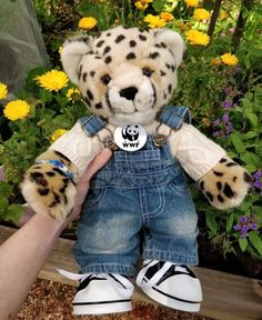 a stuffed animal that is wearing overalls and holding it's hand in front of some flowers