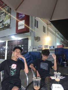 two boys sitting at an outdoor table with drinks in front of them and one holding his hand up