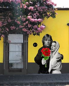 a man and woman standing next to each other in front of a yellow wall with pink flowers