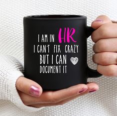 a woman holding a black coffee mug with pink writing on the side and i am in hr, i can't fix crazy but i can document it