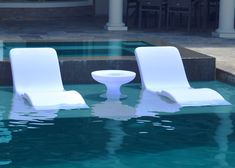 two white lawn chairs sitting in the middle of a pool next to a table and chair