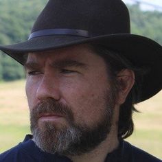 a man with a beard wearing a hat and looking off into the distance in a field