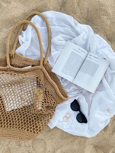 an open book and sunglasses on the sand with a netted bag next to it