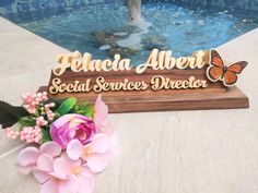 a wooden sign sitting next to a pool with flowers on the ground and a fountain in the background