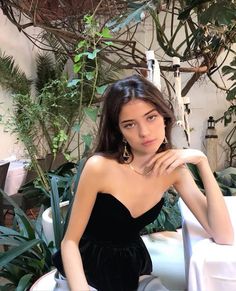 a woman in a black dress sitting at a table with potted plants behind her