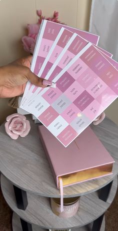 a person holding up a pink book on top of a table with flowers in the background