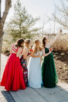 four women in dresses are standing on the sidewalk and one is pointing at something with her hand