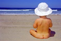 a baby sitting on the beach wearing a white hat and looking out at the ocean