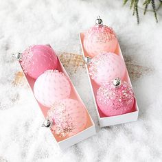 three pink and white ornaments are in a box on the snow with pine branches behind them