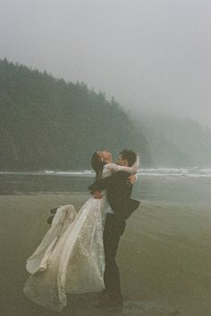 a bride and groom hugging on the beach in front of some water with trees behind them