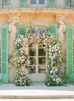 an arch covered in flowers and greenery next to a building with green shutters