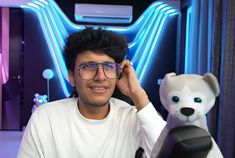 a man with glasses sitting next to a teddy bear in front of a neon sign