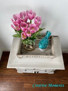 pink tulips are sitting in a white tray on a wooden floor next to a blue bird figurine