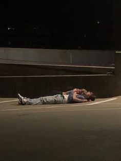 two people laying on the ground in an empty parking lot at night with their arms around each other