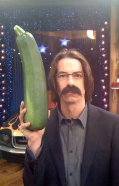 a man holding up a giant cucumber in front of his face while wearing glasses