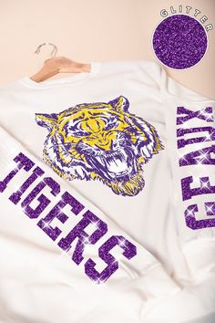 Our Glitter Geaux Tigers sweatshirt is perfect for a gameday in Tiger Stadium! This is an oversized sweatshirt that comes in sizes S/M (2-6) and M/L (8-12). This trendy long-sleeve shirt features a bold tiger graphic in vibrant yellow and purple on the front, making it a standout piece for any casual wardrobe. The shirt also showcases "Tigers" in glittery purple lettering along the sleeve, adding a touch of sparkle and school spirit. Perfect for game days, casual outings, or lounging in style, t Tiger Stadium, Tiger Graphic, Geaux Tigers, Headband Jewelry, Yellow And Purple, Over Sized, Oversized Sweatshirt, Dress Romper, Next Week