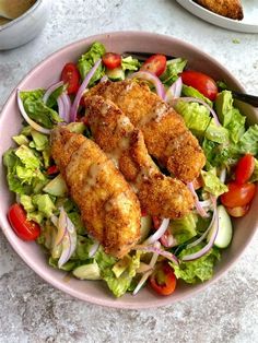 a pink bowl filled with salad and chicken on top of a white tablecloth next to two plates of food