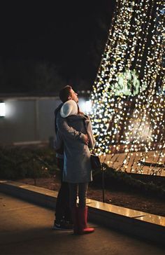 two people standing next to each other in front of a christmas tree with lights on it