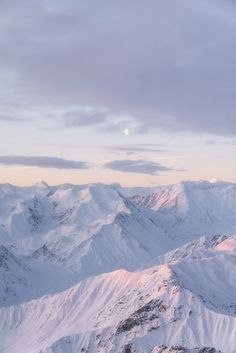 snow covered mountains under a cloudy sky with the moon visible in the distance above them