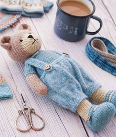 a knitted teddy bear laying on the ground next to some knitting supplies and a cup of coffee