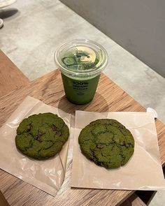 two chocolate chip cookies and a green drink on a wooden table with plastic wrappers