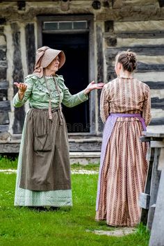 two women dressed in old fashioned clothing standing next to each other on the grass outside of a log cabin