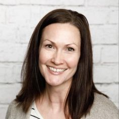 a woman with long brown hair smiling at the camera, in front of a white brick wall