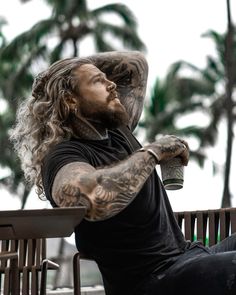 a man with long hair and tattoos sitting on a bench holding a cup in his hand