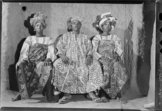 three african women in traditional dress sitting next to each other on a stone wall, looking at the camera