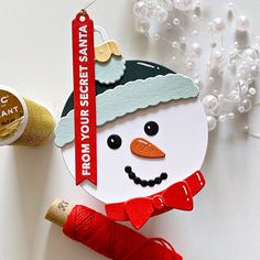 a paper snowman ornament with a red ribbon on it next to some spools of thread