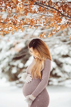 a pregnant woman standing in the snow under a tree with her hands on her belly