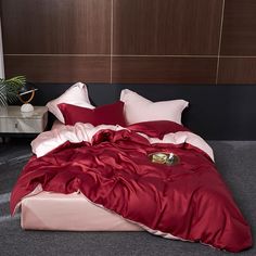 a bed with red sheets and pillows in a room next to a plant on the floor