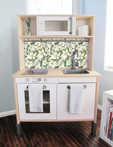 a toy kitchen with sink, microwave and cabinets in the corner by a window that has flowers on it