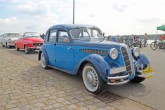 an old blue car is parked on the side of the road as people ride bikes in the background