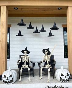 two skeletons sitting on chairs in front of a white wall with black hats and pumpkins