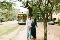 a man and woman standing next to each other near a tree with a trolley car in the background