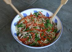 a bowl filled with noodles and veggies on top of a stone floor next to two wooden spoons