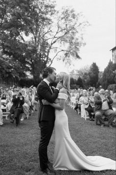 a bride and groom kissing in front of an audience at their outdoor wedding ceremony, black and white photograph