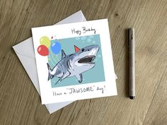 a birthday card featuring a shark with balloons on it and a pen next to it