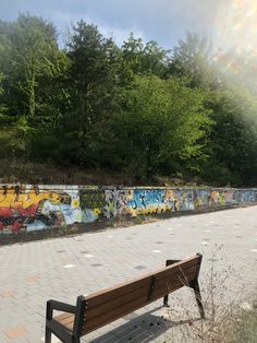 a wooden bench sitting in front of a wall covered with graffitti and trees