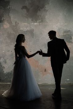 a man and woman standing next to each other in front of a dark background holding hands