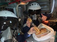 two people wearing helmets are eating sandwiches in a space station setting, while one person is holding a knife and fork