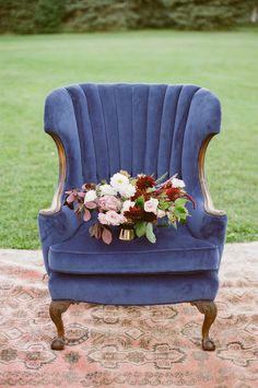 a blue velvet chair with flowers on it in front of a grassy field and trees