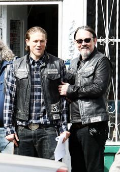 two men standing next to each other in front of a building wearing leather jackets and jeans