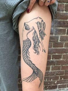 a woman's leg with a mermaid tattoo on it and a brick wall behind her