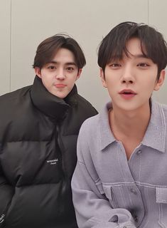 two young men sitting next to each other in front of a white wall and one is wearing a black jacket