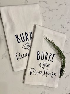 two white towels with black lettering on them sitting on a counter top next to a plant